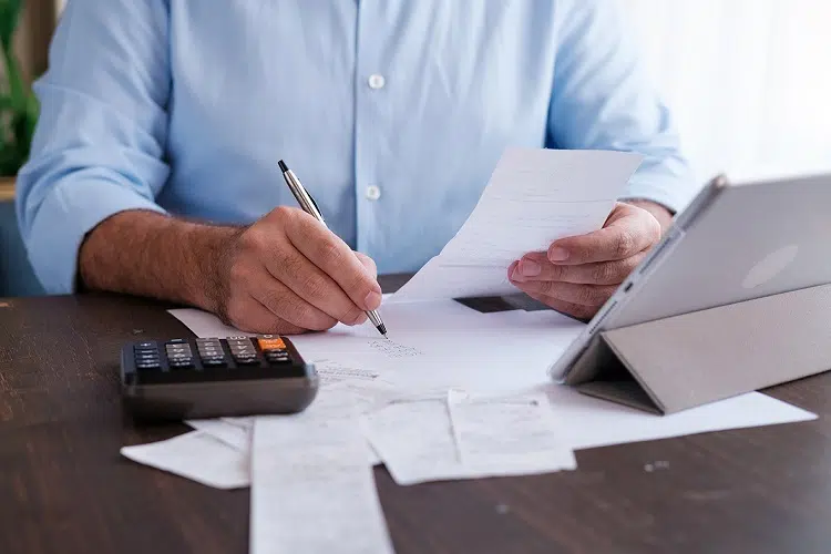 Cálculo de créditos de PIS e COFINS Homem verificando contas escrevendo em uma mesa com papeis e calculadora.