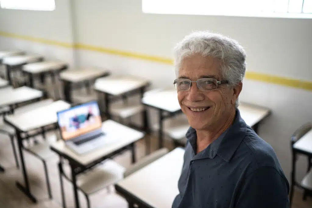 Professor homem velho de cabelos brancos sorrindo enquanto ministra aula.